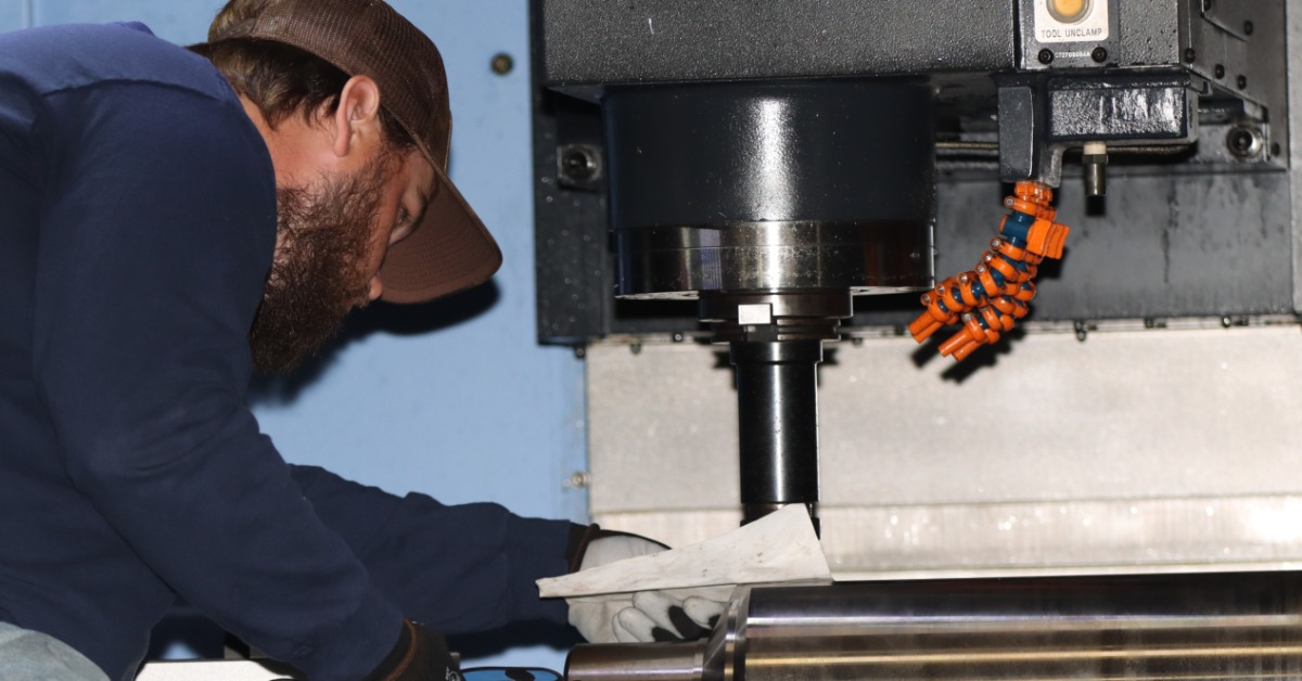 A man wearing a cap and gloves leaning toward a large machine manufacturing augers with metal rollers and orange coolant nozzles.